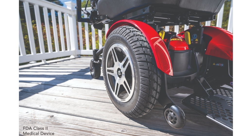 Close-up view of the rear wheels of a red Jazzy Elite HD Power Wheelchair on a wooden deck, with a white railing and green foliage in the background.
