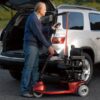 Man holding remote for Curb Sider lift in back of silver vehicle attached to red three wheel scooter