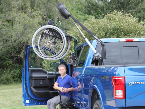 Young Man in front transfer seat in blue pickup truck pushing remote with wheelchair hoisted up attached to Out Rider lift in back bed of truck