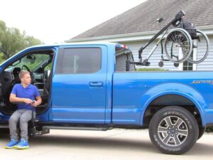 Young Man in transfer seat of blue pickup truck looking at wheelchair attached to Out Rider lift hoisted up over back bed of pickup truck