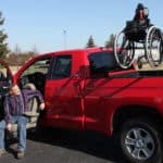 man in turning seat in red pickup truck with out-rider hoist lift in back of truck bed