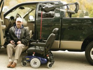 Man in turn-out transfer seat next to power wheelchair attached to Out Rider lift in back of dark green pickup truck
