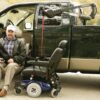 man in turn-out transfer seat next to power chair attached to lift in back bed of dark green pickup truck