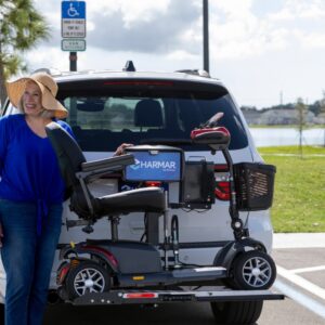 A woman stands next to an SUV with a Harmar Universal Scooter Lift (AL100) securing her mobility scooter on the back, parked in a disabled parking space near a lake.
