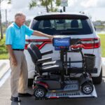 An older man in a blue shirt loads a mobility scooter onto a Harmar Universal Scooter Lift (AL100) attached to the back of an SUV in a parking lot.