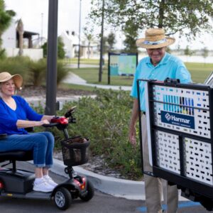 An older woman on a mobility scooter and an older man in a straw hat stand next to a Harmar Universal Scooter Lift (AL100) in a parking lot.