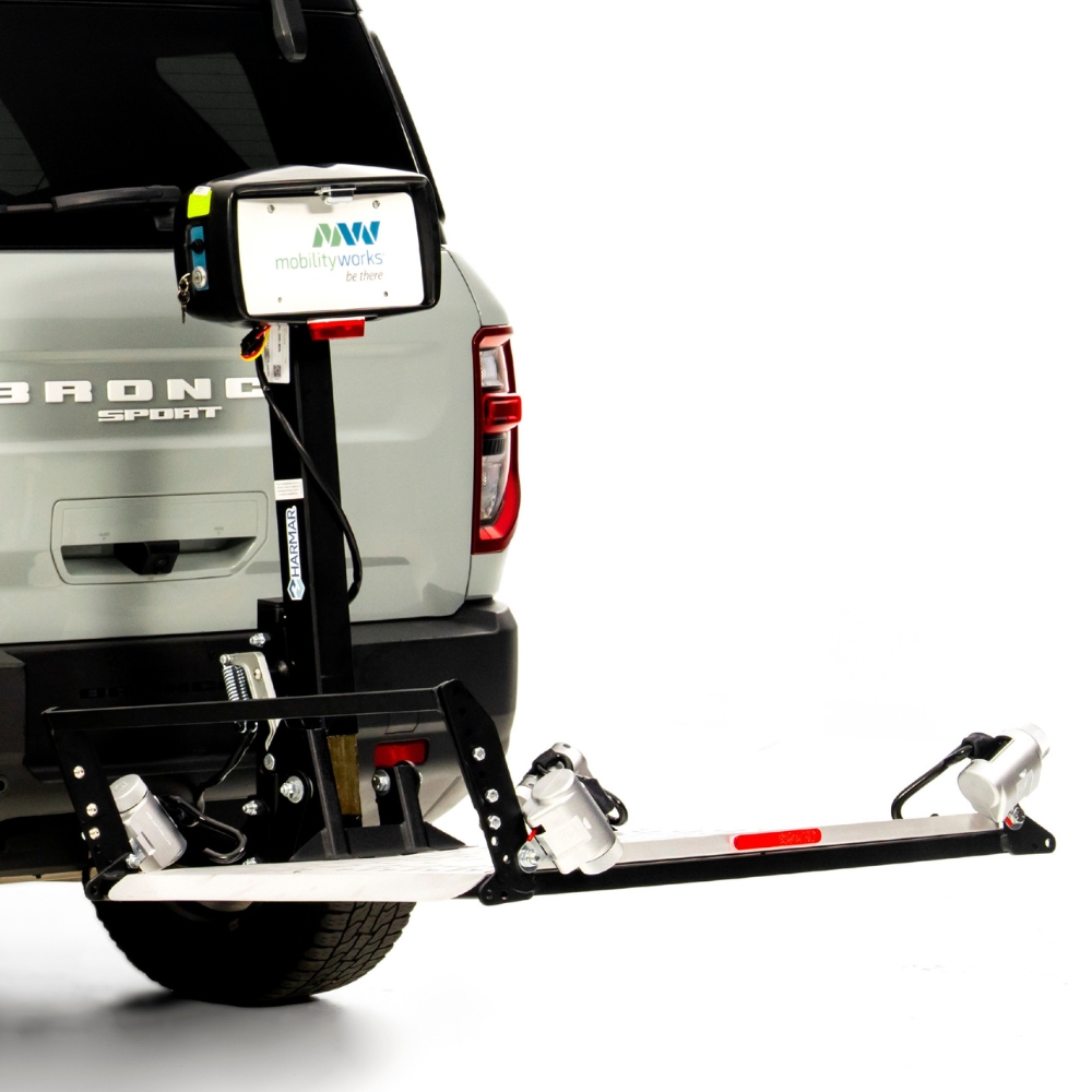A Harmar Fusion Lift (AL300-MW) is mounted on the rear of an SUV for transporting mobility devices, featuring a control box labeled with the MobilityWorks logo.