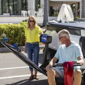 A woman assists a man on a mobility scooter as the Harmar Extra Large Fusion Lift (AL301XL) operates beside their pickup truck in a parking lot.