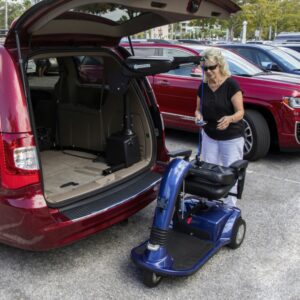 A woman uses a Harmar Premium Inside Lift (AL435) to load a blue mobility scooter into the back of a red minivan in a parking lot.