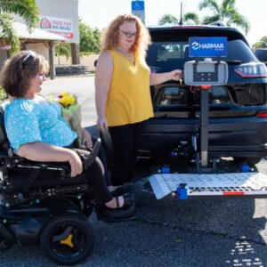 Two women are by an SUV with a Harmar Universal Powerchair Lift (AL500). One sits in a power wheelchair holding flowers, while the other operates the AL500 lift in a parking lot.