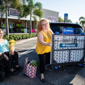 Two women load groceries into a car in a parking lot; one is seated in a power wheelchair while the other operates the Harmar Universal Powerchair Lift (AL500).