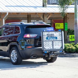 A black SUV with a Harmar Universal Powerchair Lift (AL500) attached to its rear is parked in front of a store displaying yellow sale signs in the windows.
