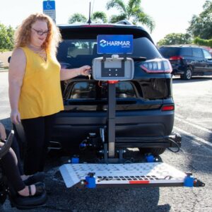 A woman stands beside a parked SUV equipped with the Harmar Universal Powerchair Lift (AL500), preparing to operate it in an accessible parking lot.