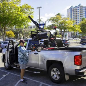 A man unloads a foldable mobility scooter from the bed of his silver pickup truck in a sunny parking lot using the Harmar 150 Speed Crew Cab Lift (AL815CC).