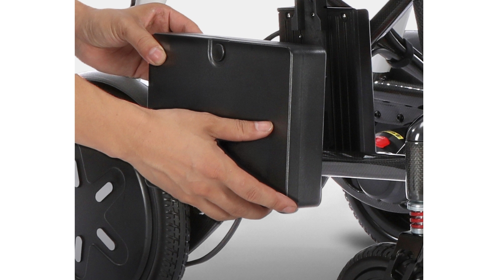 A person’s hands remove a rectangular black battery from the side of a Modo Breeze mobility scooter, near its wheel and suspension.