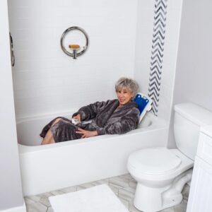 An older woman in a gray robe relaxes in a bathtub, smiling as she uses the BathLyft Power Bathtub Lift remote. The serene space, with white tiled wall and nearby toilet, suits her comfortable bathing experience.