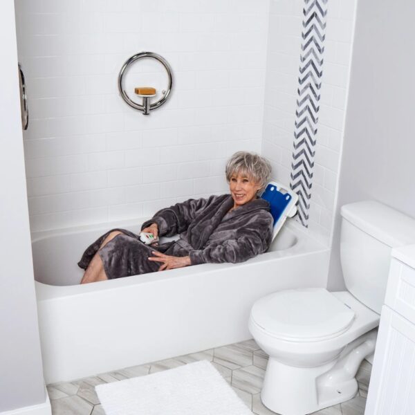An older woman in a gray robe relaxes in a bathtub, smiling as she uses the BathLyft Power Bathtub Lift remote. The serene space, with white tiled wall and nearby toilet, suits her comfortable bathing experience.