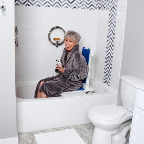 An older woman in a bathrobe sits on a BathLyft Power Bathtub Lift in a white bathroom, holding a remote and smiling—enjoying comfort and accessibility.