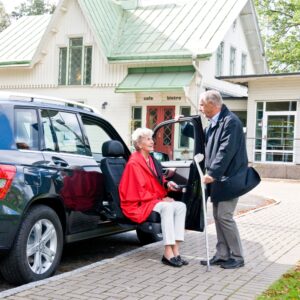 An older woman sits on a BraunAbility Turny Evo swivel car seat next to an SUV, while an older man with a cane stands nearby. In the background, there is a cafe and bistro building.