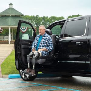A man with prosthetic legs sits on a BraunAbility Turny Evo seat extended outside a black car parked in a handicap spot near a park with a gazebo in the background.