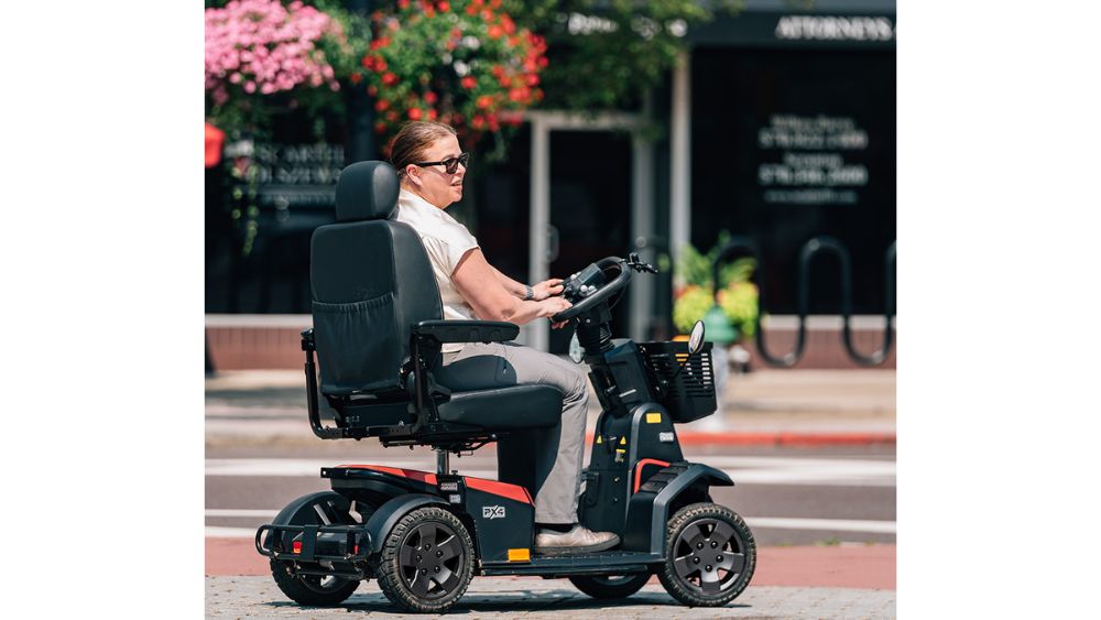 A person wearing sunglasses is riding a black and red PX4 4-Wheel Scooter by Pride Mobility along a city sidewalk, with shops and flowers visible in the background.