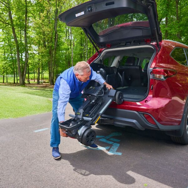 An older man loads a folded Buzzaround CarryOn HD 4-Wheel Scooter into the trunk of a red SUV parked in a handicapped space near a wooded area.