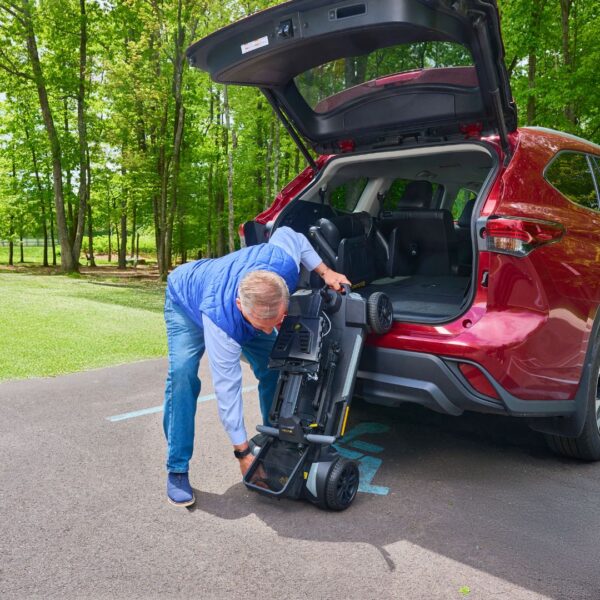 A man loads a folded Buzzaround CarryOn HD 4-Wheel Scooter into the trunk of a red SUV parked in an accessible spot near a wooded area.