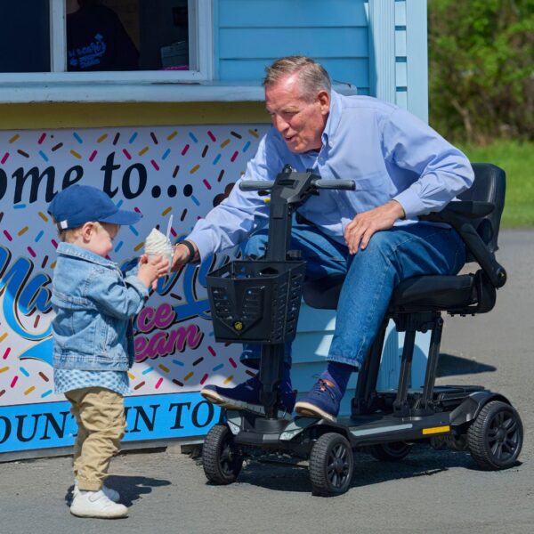 An elderly man on a Buzzaround CarryOn HD 4-Wheel Scooter receives an ice cream cone from a young boy outside an ice cream stand.