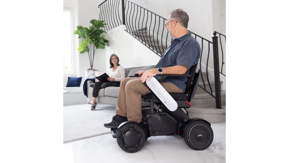 A man in a C2 Model - Portable Electric Wheelchair faces a woman sitting on a sofa in a modern living room with white walls and a staircase in the background.