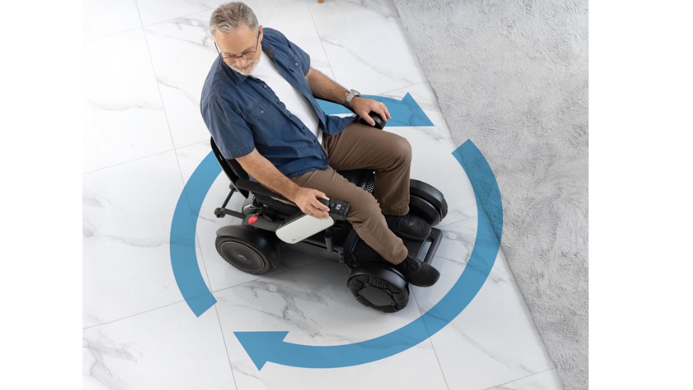 A man operates the C2 Model - Portable Electric Wheelchair on a tiled floor, demonstrating its impressive rotation capability as blue arrows highlight its ability to turn in place.