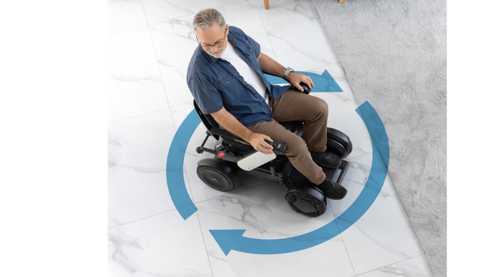 A man uses the WHILL Model C2 High Back motorized wheelchair to turn in place on a tiled floor, with blue arrows indicating the wheelchair’s rotation.