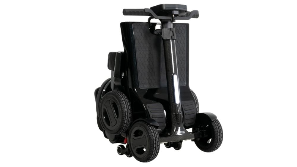 A Go Go™ Carbon 4-Wheel Scooter in black is shown folded upright, highlighting its compact and portable design with visible wheels and handlebars.
