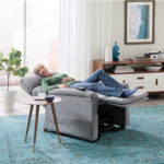 A woman relaxes on a Cloud Lift Chair w/ MaxiComfort with her feet up, beside a small round side table in a bright, modern living room.