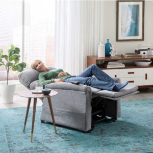 A woman relaxes on a Cloud Lift Chair w/ MaxiComfort with her feet up, beside a small round side table in a bright, modern living room.