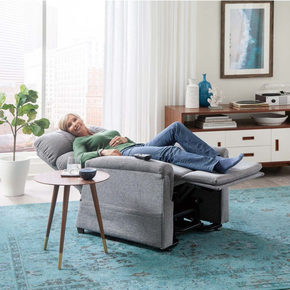 A woman relaxes on a Cloud Lift Chair w/ MaxiComfort with her feet up, beside a small round side table in a bright, modern living room.