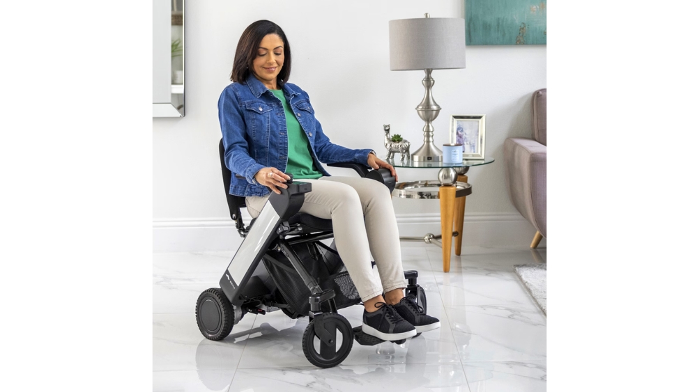 A woman sits in a WHILL Model F motorized wheelchair in a living room with white floors, wearing a denim jacket, green shirt, and beige pants. Nearby, a lamp and framed photo rest on a side table.