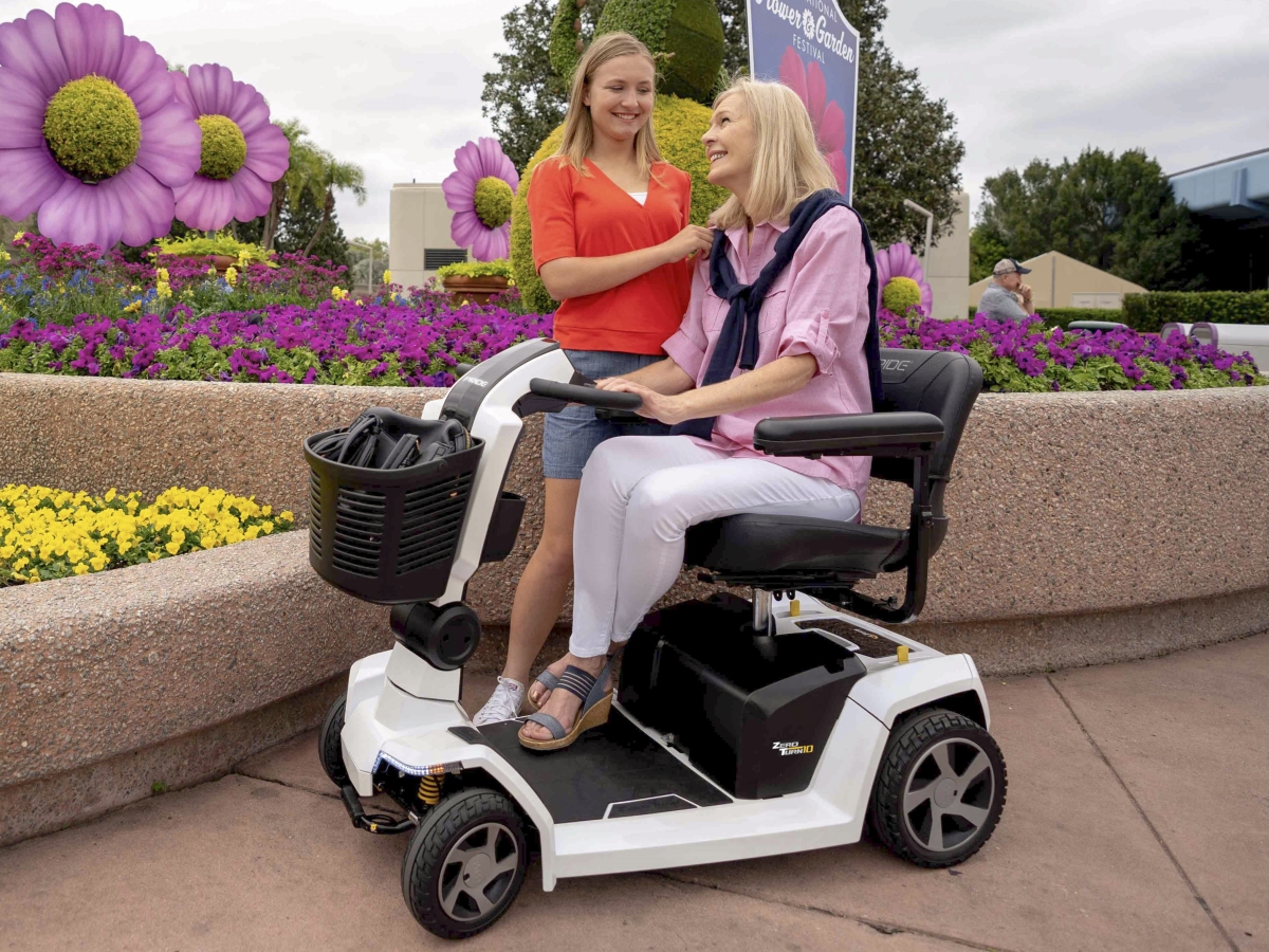 A woman in a mobility scooter smiles and talks with another woman standing beside her. They are in a garden setting with large floral decorations in the background.