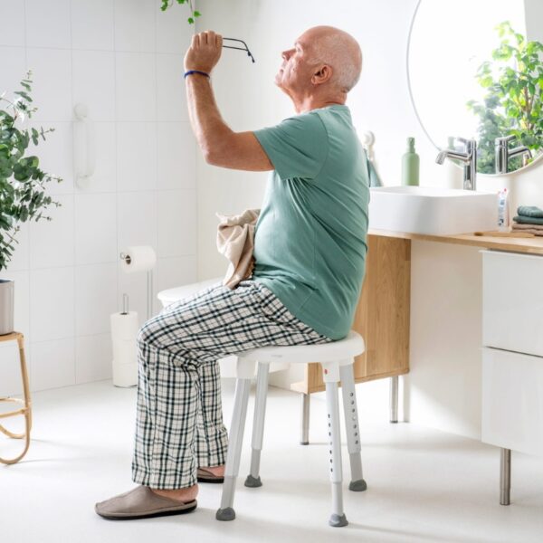 An older man in pajamas sits on an Etac Easy Shower Stool in the bathroom, holding his glasses up and examining them with a towel in hand.