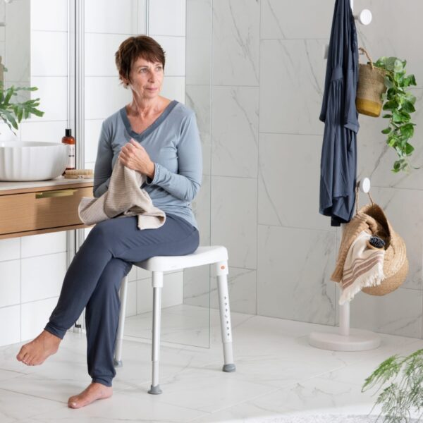 An older woman in comfy clothes sits on an Etac Edge Shower Stool in a modern bathroom, holding a towel, with toiletries and hanging items visible in the background.