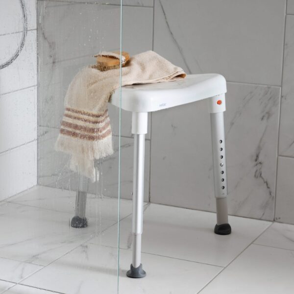 The Etac Edge Shower Stool with adjustable legs stands in a tiled shower, holding a beige towel and bar of soap on its white seat.