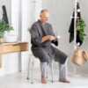 A man in a gray robe sits on the Etac Smart Shower Stool in a modern bathroom, holding and cleaning his glasses.