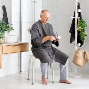 A man in a gray robe sits on the Etac Smart Shower Stool in a modern bathroom, holding and cleaning his glasses.