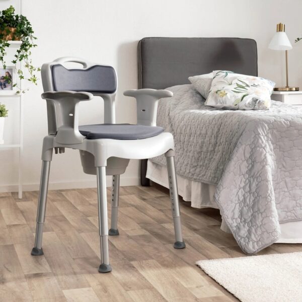 A gray adjustable bedside commode chair with armrests is placed next to a neatly made bed in a bright, tidy bedroom.