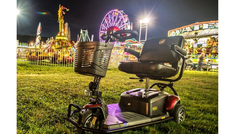 A BuzzAround LX 3-Wheel Scooter is parked on grass at night in front of a brightly lit carnival, with rides such as a Ferris wheel and a food stand visible in the background.