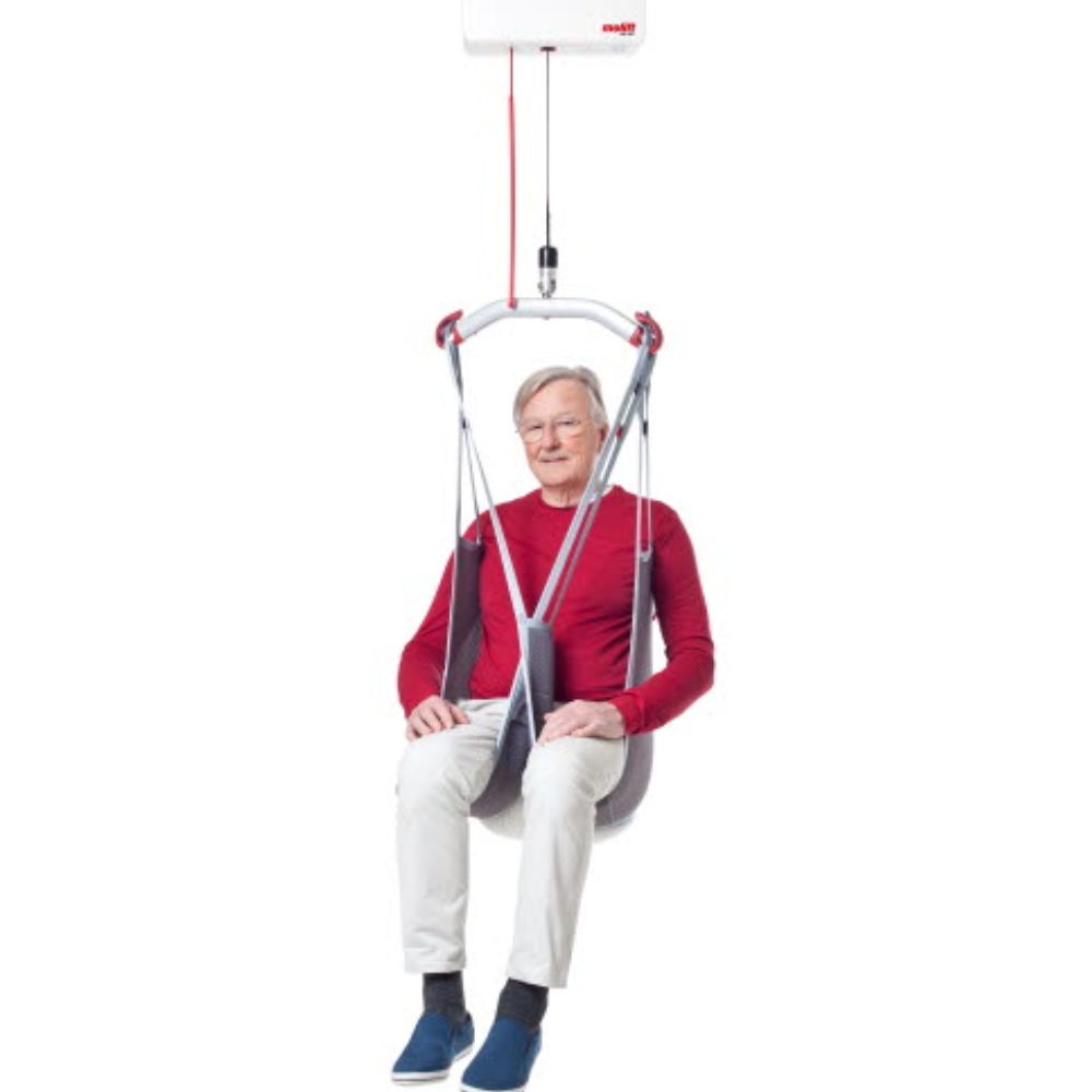 An older man sits in an Evosling Lowback Net attached to a ceiling lift system, demonstrating patient transfer equipment with the Nova Stand-Up for enhanced mobility support.