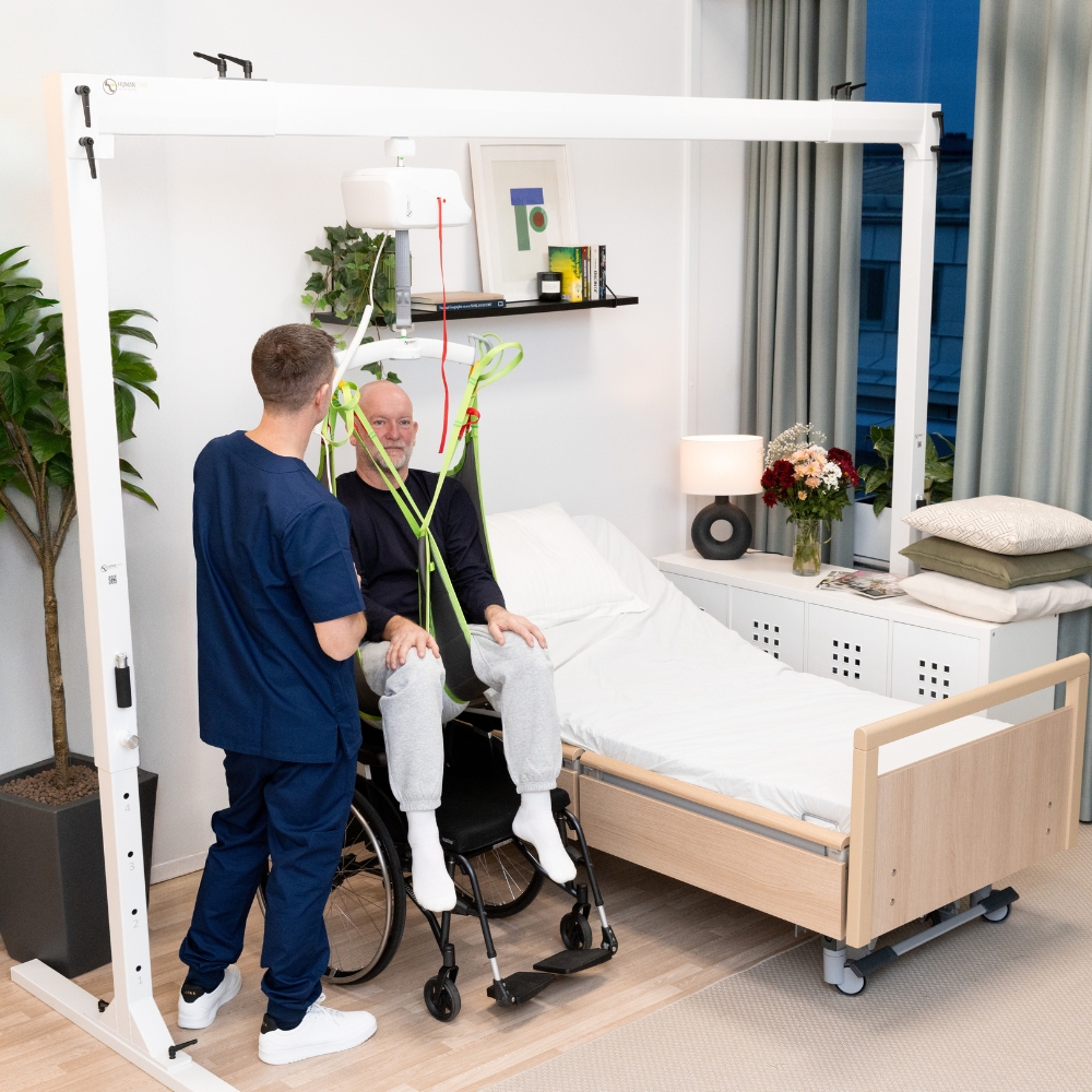 A healthcare worker assists an elderly man in a wheelchair with a ceiling lift transfer using the FlexiRail Freestanding Gantry Lift, positioned next to a hospital bed in a modern, well-lit room.