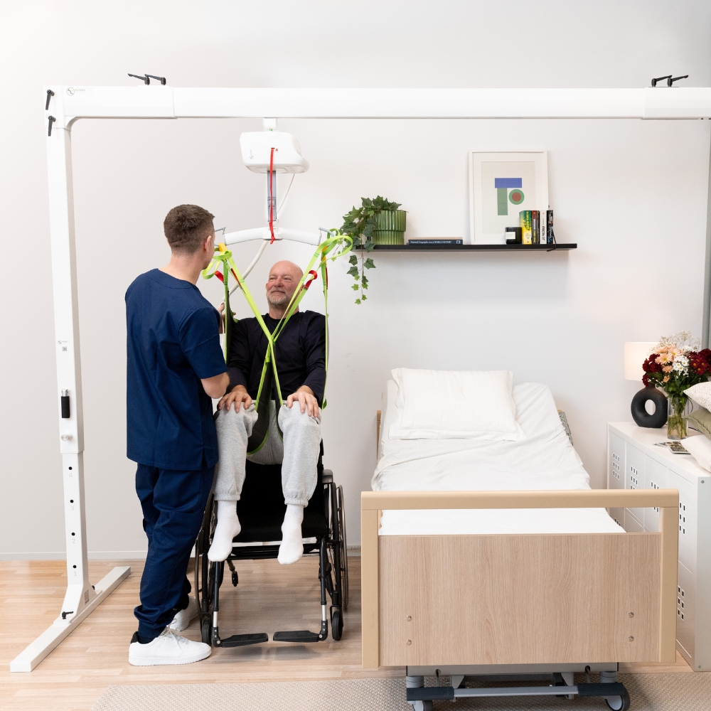 A nurse assists a seated patient in a transfer sling lift connected to a FlexiRail Freestanding Gantry Lift next to a hospital bed in a modern, bright patient room.