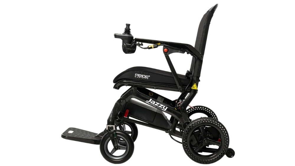 Jazzy® Ultra Light Travel Power Wheelchair in black, featuring a joystick controller on the armrest, padded seat, footrest, and perforated wheels, shown from a side view.