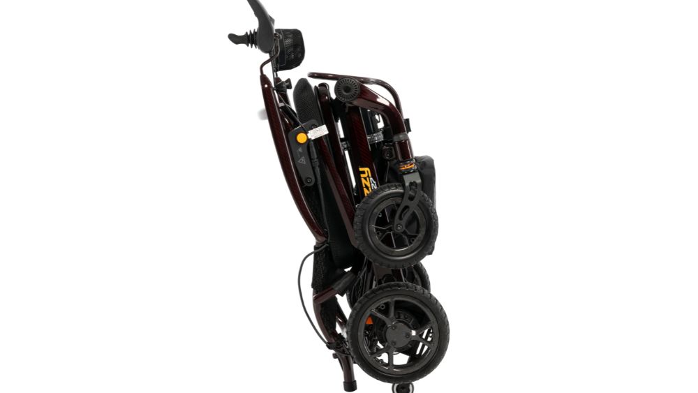 A compact, folded Jazzy Carbon 27 Power Wheelchair in black and red is standing upright on its wheels, viewed from the side against a plain white background.