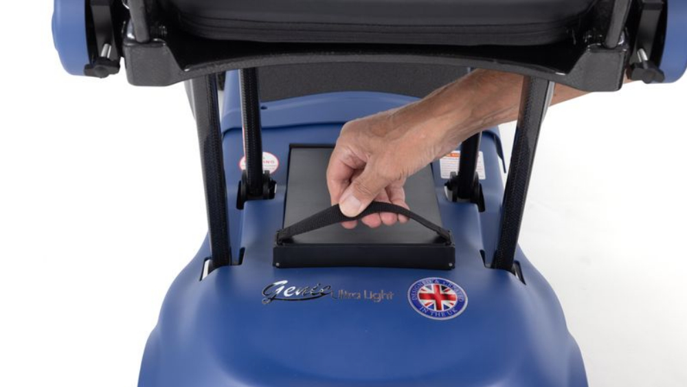A hand pulls a fabric strap to lift a black battery pack from the base of a Monarch Mobility Genie Ultralight mobility scooter.